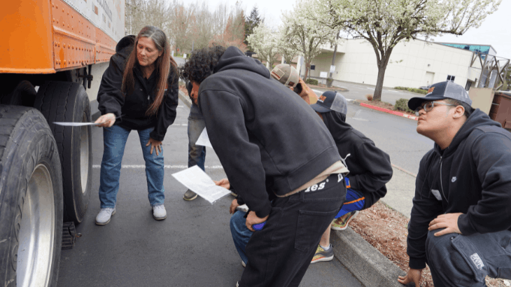 Nicole pointing with a stick at semi truck wheel, students looking at wheel