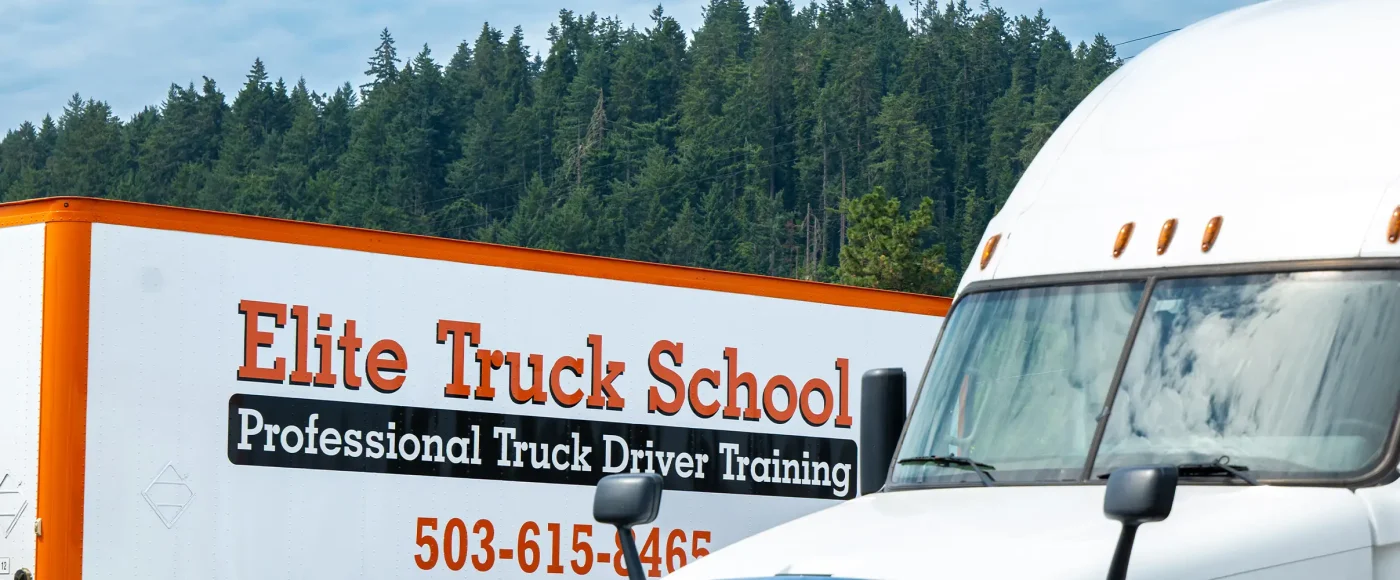 Close-up of an Elite Truck School trailer beside a white semi-truck cab, with a forested hillside behind them.