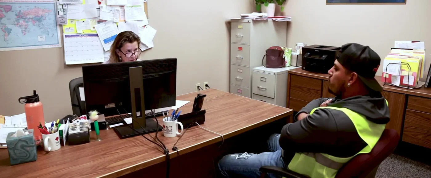 A prospective student in a safety vest meeting with an Elite Truck School admissions staff member across a desk in an office