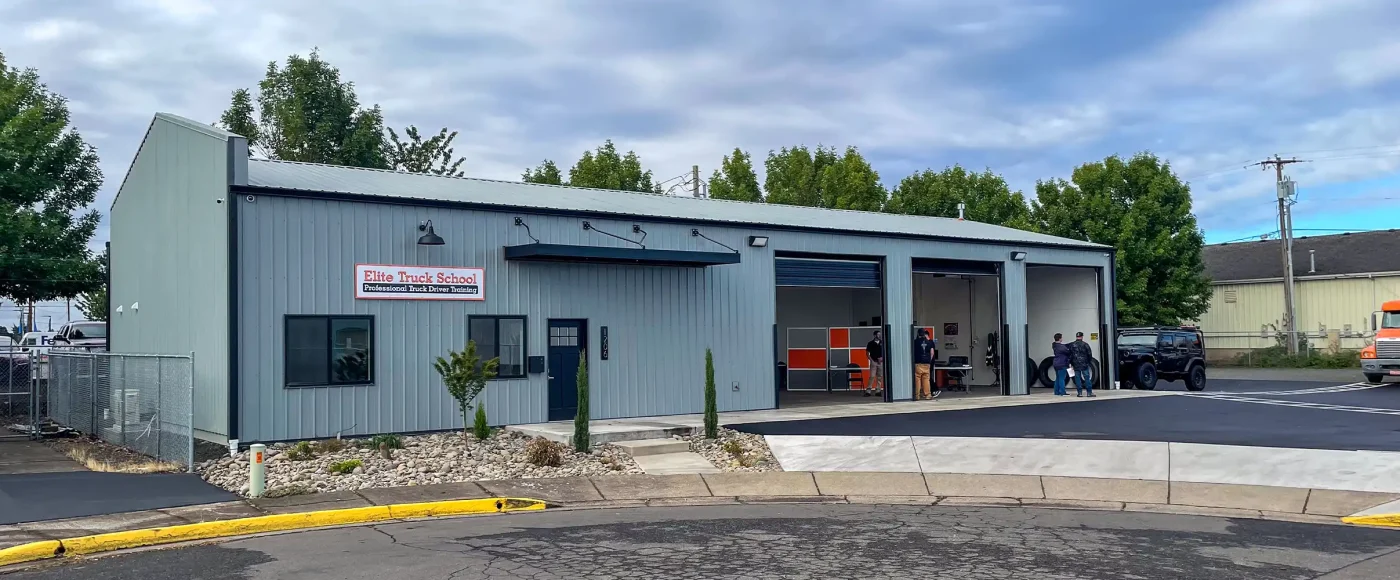 Exterior of the Albany location, a gray metal building with open garage bays where staff and students gather, a signed entrance, and a semi-truck parked nearby.