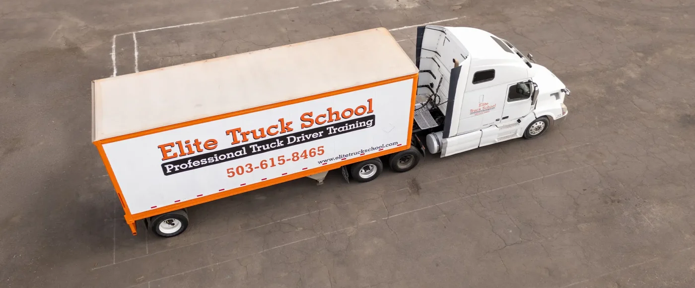 Aerial view of an Elite Truck School semi-truck with a white and orange branded trailer parked on a paved lot.