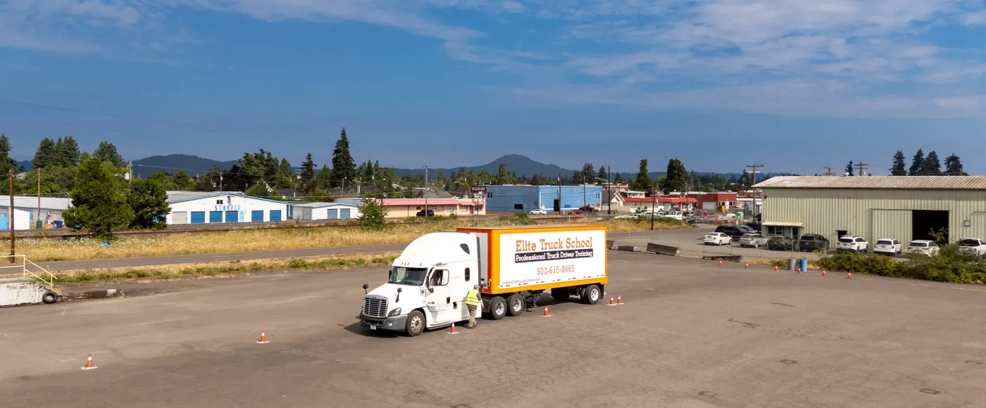 Elite semi-truck with an orange and white trailer parked on a cone-marked training lot, with a student in a safety vest beside it