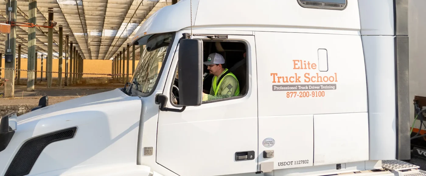 A driver in a safety vest and cap seated in the cab of a white Elite Truck School semi-truck, viewed from the side through the open window.