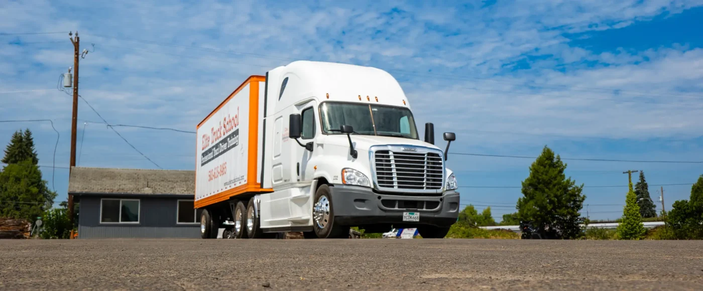 An Elite Truck School semi-truck with an orange and white trailer parked on pavement, shot from a low angle with buildings and trees behind it.