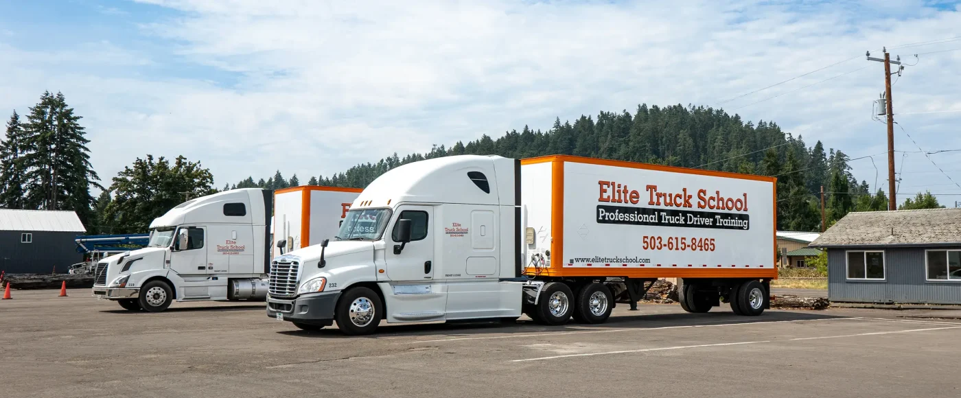 Two Elite Truck School semi-trucks with orange and white trailers parked in a paved lot, with trees and buildings in the background.