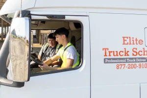 An Elite Truck School instructor and a student in safety vests seated together in the cab of a branded semi-truck, with the student at the wheel