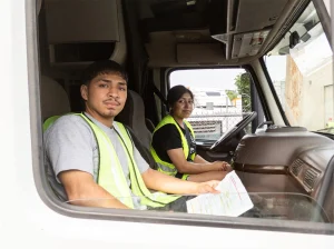 Two CDL students in safety vests seated in the cab of a semi-truck with an air brakes study sheet, viewed through the passenger window
