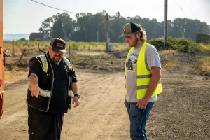 An instructor in a safety vest talking with a student in a safety vest on a dirt lot, with fields and trees in the background