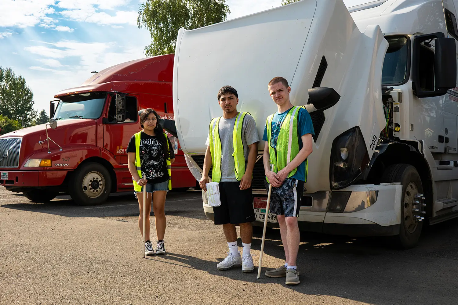 Three CDL students in safety vests holding inspection sticks standing between a red and a white Elite Truck School semi-truck in a training lot