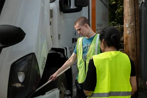 A CDL student in a safety vest using a tire thumper to inspect the front tire of a white semi-truck during a pre-trip inspection, with another student looking on