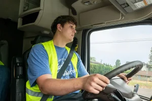 A young CDL student in a safety vest gripping the steering wheel inside the cab of a semi-truck, looking ahead through the windshield