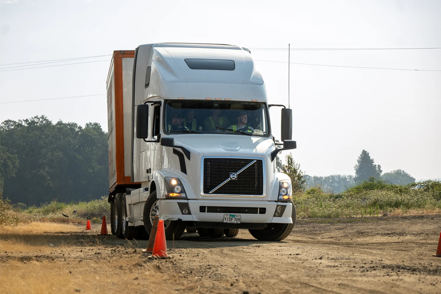 A CDL permit student drives a white semi truck through an orange cone course on a dirt training range while an instructor and other students in safety vests ride along in the cab.