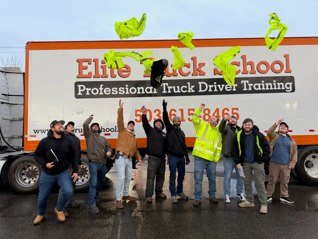 A group of smiling Elite Truck School graduates celebrate by tossing their neon yellow safety vests into the air in front of a school trailer