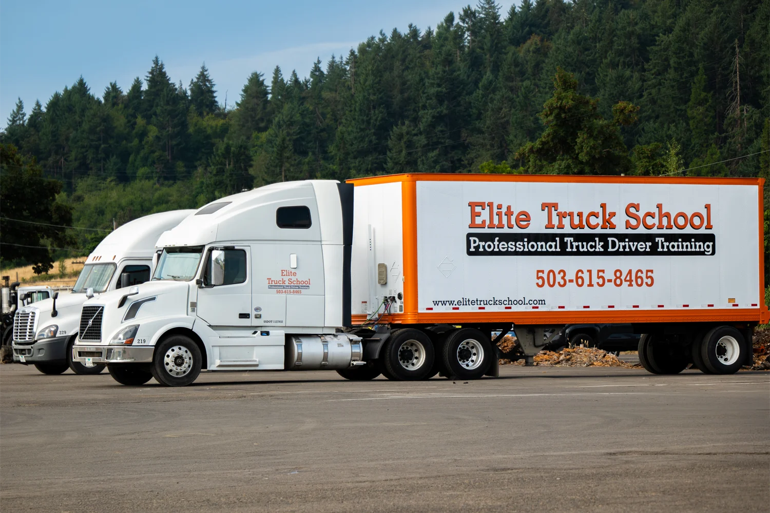 Two Elite Truck School semi trucks parked side-by-side in a paved training yard with spruce trees
